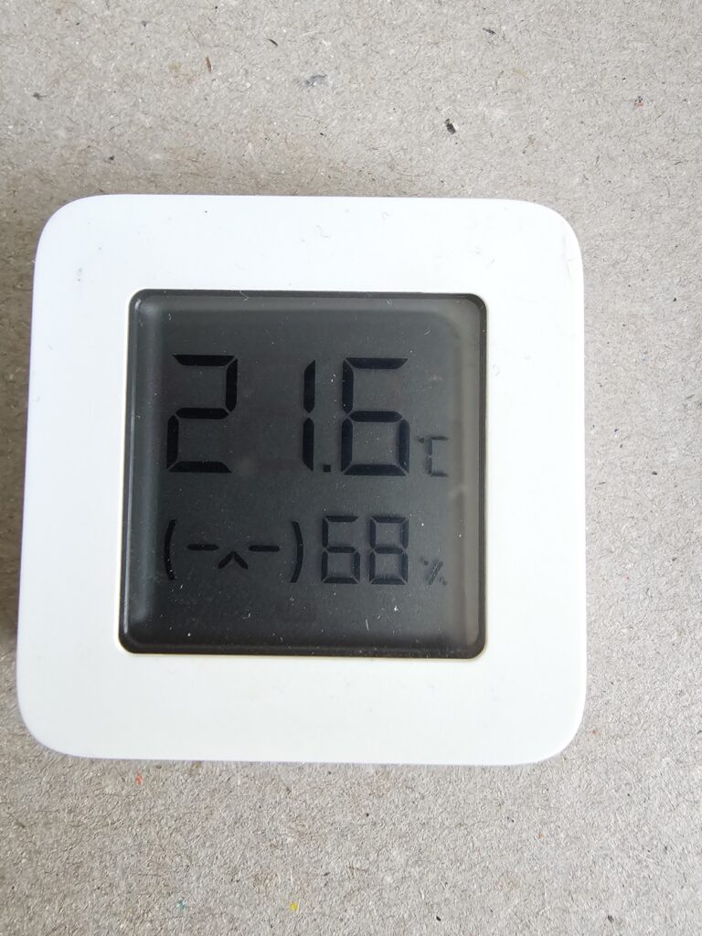 A photo of a white, square thermometer with rounded corners, with a prominent monochrome non-backlit LCD screen. In big 7-segment digits it display a temperature of 21.6°C, on the bottom it displays an ASCII art sad face to the left and a relative humidity of 68% to the right.